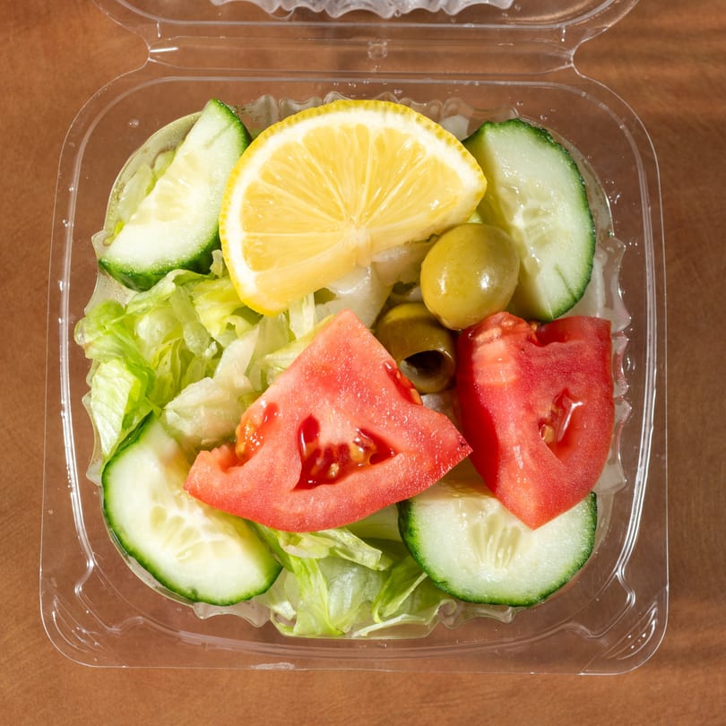 Zaytoon's fresh side salad with lettuce, cucumber, tomato, olives, and a lemon wedge in a plastic container.
