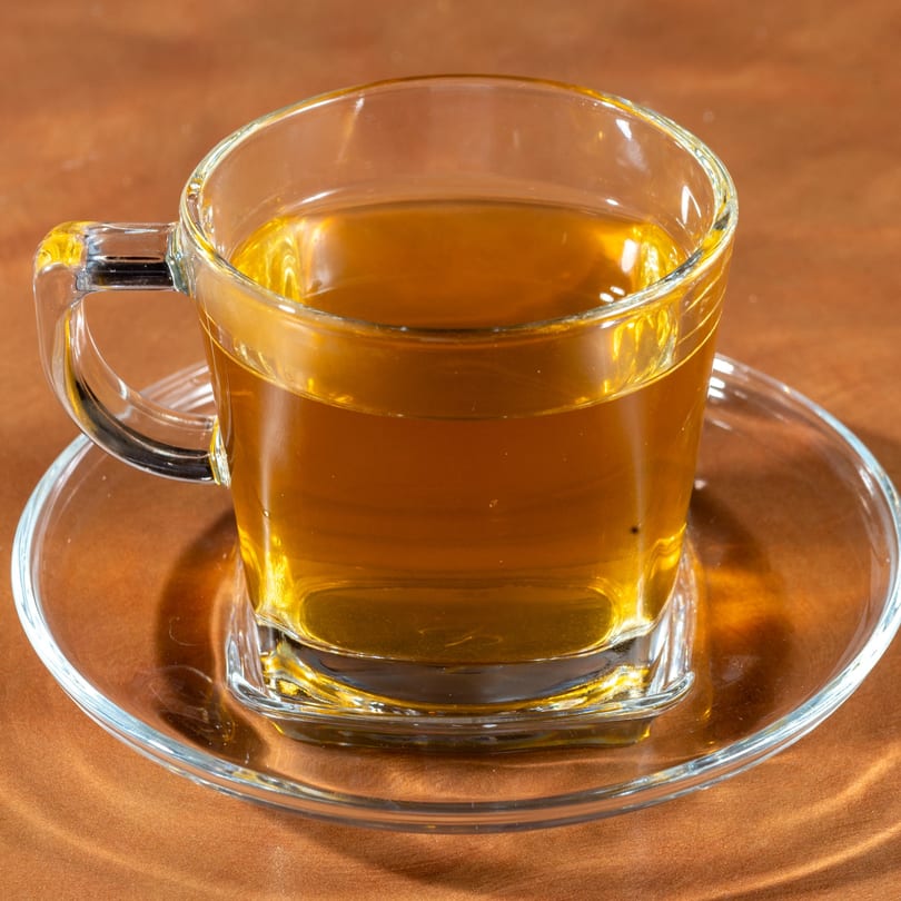 Clear glass cup of golden Afghan tea on a matching saucer, against a warm brown background.