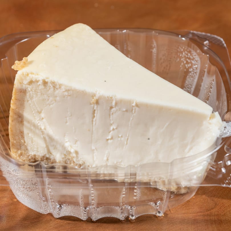 A slice of cheesecake with a graham cracker crust in a clear plastic container.