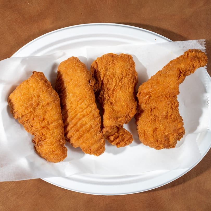 Four crispy, golden-brown fried chicken tenders on white parchment paper, served on a white plate.
